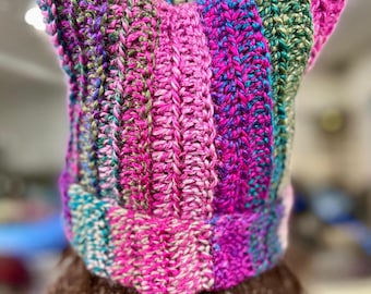 May include: A hand-knitted beanie with a cat-ear silhouette, featuring a vibrant vertical striped pattern in shades of magenta, bright pink, teal, forest green, and purple. The textured yarn creates a ribbed effect throughout the entire hat.