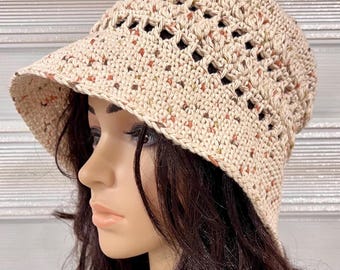 May include: A beige crochet bucket hat featuring a speckled pattern with small flecks of orange, green, and brown throughout the yarn. The hat has a wide, downward-sloping brim and a decorative open-stitch band around the crown area.