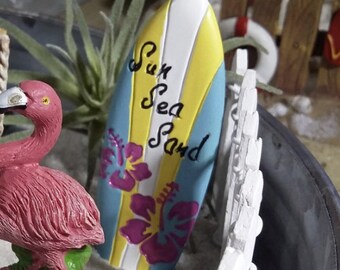 May include: A miniature pink flamingo figurine stands on a small gray rock, next to a white picket fence and a blue, yellow, and white surfboard with the words "Sun Sea Sand" written on it. The scene is set in a miniature garden with white sand.
