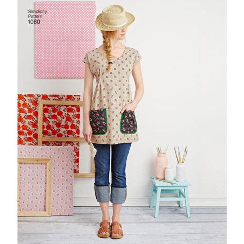 May include: A woman in a beige tunic with red floral patterns and two floral-patterned pockets. She wears blue jeans with turned-up cuffs, brown sandals, and a straw hat. The image includes framed artwork and the text "Simplicity Pattern 1080".