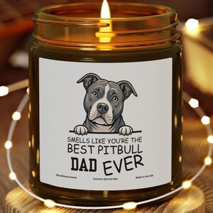 Peut inclure: Une bougie allumée dans un bocal en verre avec une étiquette indiquant "Smells Like You're The Best Pitbull Dad Ever". L'étiquette présente un pitbull de dessin animé. La bougie repose sur un dessous de verre en bois.