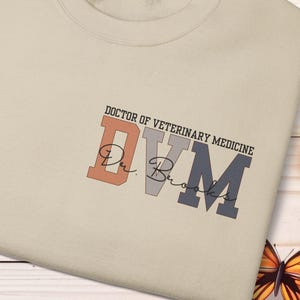 May include: A beige sweatshirt with the text "DOCTOR OF VETERINARY MEDICINE" and "DVM" in a mix of colors. The sweatshirt has a crew neck and is decorated with butterflies and flowers.