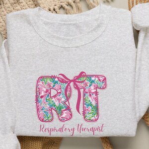 May include: A light gray crewneck sweatshirt featuring a floral-patterned graphic of the letters RT with a pink ribbon tied around them. Below the letters, the text Respiratory Therapist is printed in a matching pink script font.