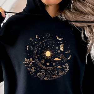 May include: Black hooded sweatshirt with a celestial design. The design features a sun, moons, stars, and floral elements in a gold color. The text "Black" and "Gildan 18500" are printed on the bottom left of the sweatshirt.