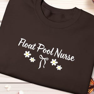May include: A dark brown t-shirt with the words "Float Pool Nurse" in white script, decorated with white daisies and a bow. The shirt is displayed on a light wood-grain surface, with butterflies and flowers.