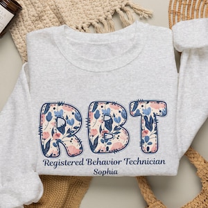 May include: A light gray crewneck sweatshirt featuring the letters RBT in a floral pattern with blue, pink, and cream tones. Below the letters, the text reads Registered Behavior Technician and the name Sophia in a dark blue font.