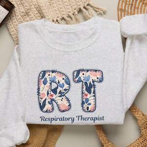 May include: A light gray crewneck sweatshirt featuring the letters RT in a floral pattern with blue and pink flowers and dark leaves. Below the letters, the text Respiratory Therapist is printed in a dark blue, simple sans-serif font.