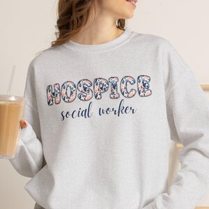 May include: A light gray crewneck sweatshirt featuring the text "HOSPICE" in a bold, floral-patterned font with blue and pink flowers, followed by the words "social worker" in a dark blue cursive script, paired with gray denim shorts.