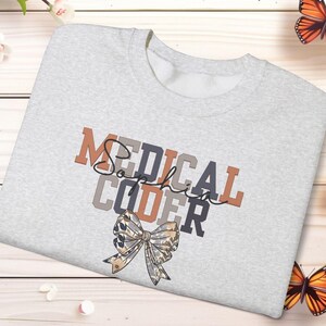 May include: A light gray sweatshirt with the words "MEDICAL CODER Sophia" in a mix of colors and fonts. A decorative bow is at the bottom. Butterflies and flowers are arranged around the sweatshirt on a light wood background.