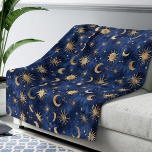 May include: A navy blue throw blanket with a celestial design featuring gold suns, moons, and stars. The blanket is draped over a white sofa, creating a cozy and decorative accent for the living room. The blanket is made of a soft material.