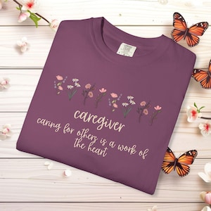 May include: A plum-colored t-shirt featuring a graphic of small wildflowers in pink, white, and green. The text reads "caregiver" and "caring for others is a work of the heart" in a light-colored script font. The shirt has a crew neckline.