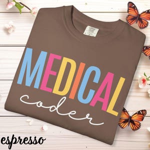 May include: Espresso-colored t-shirt with the words "MEDICAL coder" in colorful block letters. The word "MEDICAL" is in blue, yellow, and pink. The word "coder" is in white script. Butterflies and flowers are in the background.