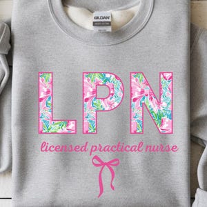May include: A Gildan sports grey sweatshirt with the letters "LPN" in a pink floral pattern. Below the letters is the text "licensed practical nurse" in pink, with a pink bow graphic. The sweatshirt is a crew neck.