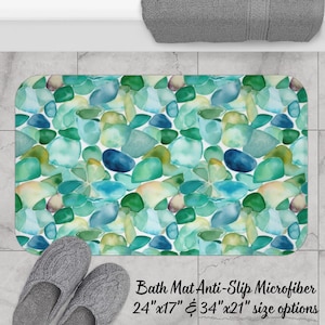 May include: A rectangular bath mat featuring a watercolor-style pattern of smooth, rounded stones in shades of teal, seafoam green, navy blue, and pale yellow. The mat is available in 24 inches by 17 inches and 34 inches by 21 inches sizes.