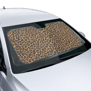 May include: A car windscreen sunshade featuring a leopard print pattern. The sunshade is in shades of brown and tan with dark brown spots. It is installed in a silver car and covers the windscreen.