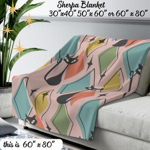 May include: A pink sherpa blanket with a mid-century modern design. The blanket features black cat silhouettes, diamonds, and starbursts in teal, yellow, and orange. The blanket measures 60" x 80".