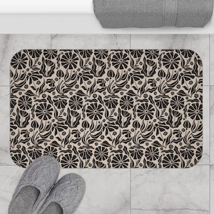 May include: A rectangular bath mat with a floral pattern in black and beige. The design features stylized flowers and leaves. Gray slippers are at the bottom of the mat. A gray towel is at the top.