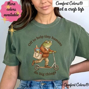 May include: Green Comfort Colors t-shirt with a frog graphic riding a scooter and carrying a tote bag. The text reads "Off to help tiny humans do big things." The shirt also has the text "More colors available" and "not a crop top."