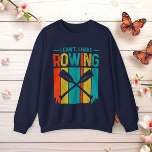 Puede incluir: Sudadera azul marino con el texto "I can't, I have Rowing" en letras de bloque coloridas. El diseño incluye remos cruzados y rayas verticales en naranja, amarillo y turquesa. Una prenda casual y deportiva.