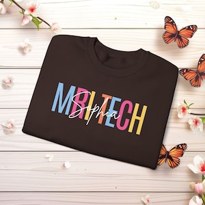 Może przedstawiać: Ciemnobrązowa koszulka z napisem "MRI TECH Sophia" w kolorowych blokowych literach i kursywą. Koszulka jest prezentowana na jasnej drewnianej powierzchni z motylami i kwiatami.
