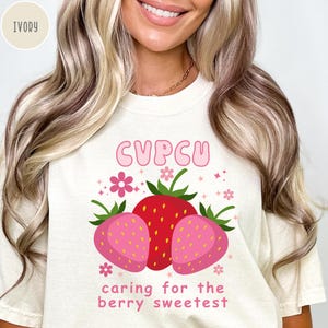 May include: Ivory-colored t-shirt featuring a graphic of three strawberries. The central strawberry is red, flanked by two pink strawberries. The text "CUPCU" is above the strawberries, with "caring for the berry sweetest" below.