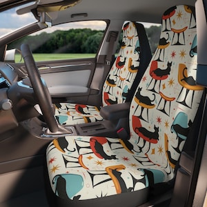 May include: Car seat covers with a mid-century modern design featuring black cat silhouettes on colorful chairs, set against a cream background with starburst accents. The covers are visible inside a car, with the steering wheel and dashboard in view.