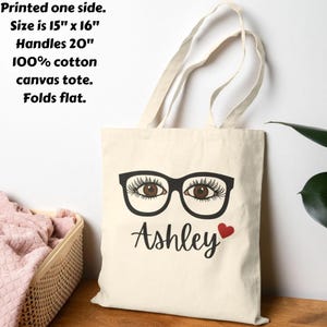 May include: Cream-colored canvas tote bag with black eyeglasses graphic and the name "Ashley" with a red heart. The bag is 15" x 16" with 20" handles and made of 100% cotton. The tote folds flat.