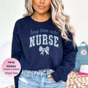 Könnte beinhalten: Marineblaues Sweatshirt mit dem Schriftzug "long term care NURSE" in floralem Design. Unter dem Wort "NURSE" befindet sich eine florale Schleife. Das Sweatshirt ist von der Marke Gildan und in weiteren Farben erhältlich.