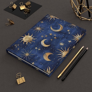 May include: A hardcover journal with a celestial design featuring gold suns, moons, and stars on a dark blue background. Includes gold and black pencils, and gold binder clips. The journal is closed and lying flat.