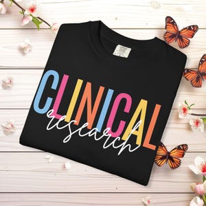 May include: Black t-shirt with the words "CLINICAL research" in colorful block letters and white cursive script. The shirt is folded on a light wood surface, decorated with butterflies and flowers.