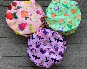 Op de afbeelding: Drie satijnen haarbonnets met verschillende patronen. Een bonnet is roze met fruit- en watermeloenontwerpen. Een andere bonnet is groen met fruit- en bladerenontwerpen. De derde bonnet is paars met een gotisch thema met vleermuizen, cupcakes en kruisontwerpen.