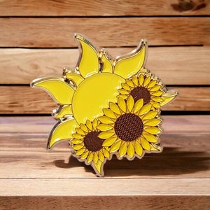 May include: A gold-toned metal pin with a stylized sun design and three sunflowers. The sun is yellow and the sunflowers have yellow petals and brown centers.