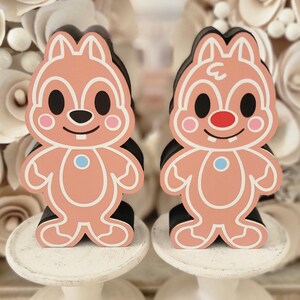 May include: Two gingerbread-style chipmunk figurines with black, pink, and white details. They stand on white pedestals in front of a backdrop of paper flowers. The text "Sandford Designs" is at the bottom.