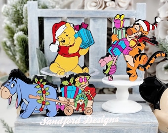 Könnte beinhalten: Holzausschnitte von Winnie-Puuh-Charakteren in Weihnachtskleidung. Puuh hält ein blaues Geschenk, Tigger trägt Geschenke, I-Aah zieht einen Wagen mit Geschenken, und Ferkel ist dabei. Die Figuren stehen auf einer grauen Bank mit einem Mickey-Mouse-Kopf.