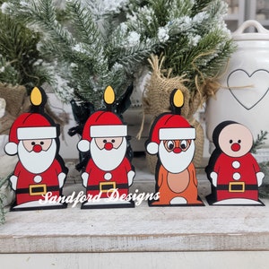 May include: Four Christmas-themed wooden cutouts. Three Santa Claus figures and one gingerbread man are depicted in red, white, black, and brown. Each figure has a yellow candle flame on top. The text "Sandford Designs" is printed on the base.