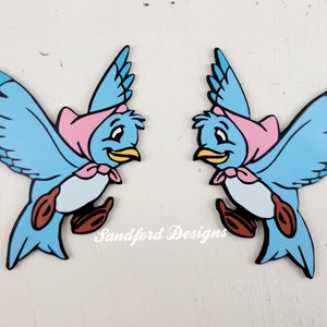 May include: Two cartoon bluebirds with black outlines, wearing pink bonnets and bows, and brown shoes. The birds have yellow beaks and are smiling. The text "Sandford Designs" is printed in white on the bottom of the image.