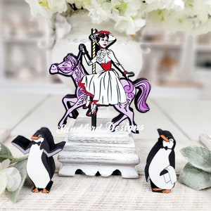 May include: A whimsical wooden carousel figure with a woman in a white dress and red accents riding a purple horse. Two penguin figures stand nearby. The figures are on a white wooden stand.