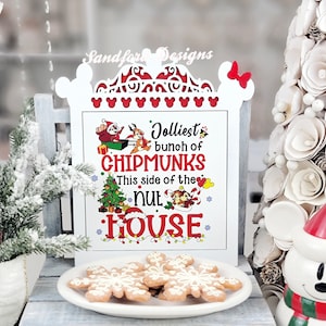 May include: A white wooden sign with a decorative top featuring Mickey Mouse silhouettes and a red bow. The sign reads "Jolliest bunch of Chipmunks, This side of the nut house." A plate of snowflake cookies sits on a small wooden stand.