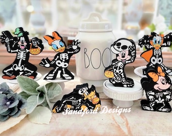 May include: A collection of Halloween-themed character figurines, including Mickey Mouse, Minnie Mouse, Donald Duck, Daisy Duck, and Goofy, all dressed as skeletons. Each figure is black with white skeleton detailing and holding a Halloween treat. A white jar with "BOO" on it is also present.