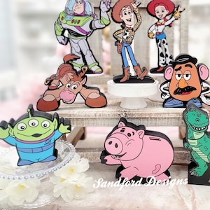 May include: A collection of colorful character cutouts from the Toy Story franchise. The figures include Woody, Buzz Lightyear, Jessie, Mr. Potato Head, Slinky Dog, Rex, Hamm, and an alien. The cutouts are displayed on a tiered wooden stand.