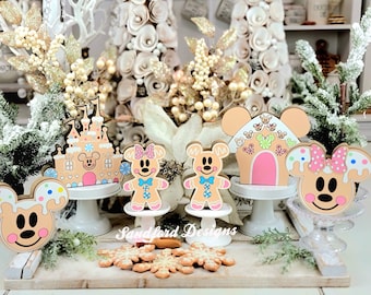 Könnte beinhalten: Weihnachtsdekorationen aus Holz mit Mickey Mouse und Minnie Mouse-Motiven. Enthält ein Lebkuchenhaus, ein Schloss und Lebkuchenmännchen. Die Dekorationen stehen auf weißen Sockeln und einem Holztablett, mit Schneeflocken-Keksen.
