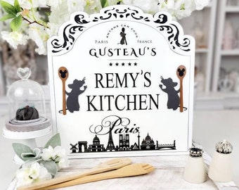 May include: A white decorative sign with the text "Remy's Kitchen" and a silhouette of the Paris skyline. The sign features illustrations of rats holding spoons and is decorated with floral accents. Includes wooden utensils and salt and pepper shakers.