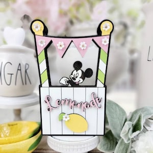May include: A decorative lemonade stand featuring Mickey Mouse. The stand has a pink and green striped awning with small white flowers. The sign reads "Lemonade" with a yellow lemon illustration. The stand is on a white pedestal.