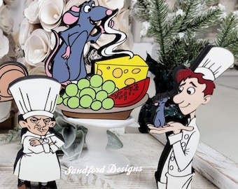 May include: Wooden cutouts of characters from the movie Ratatouille. The cutouts include a rat, a chef, cheese, grapes, and watermelon. The characters are in color and are standing on a white surface. The background includes a white floral arrangement and a small Christmas tree.