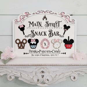 May include: A white wooden sign with pink detailing, reading "Main Street Snack Bar." The sign displays Mickey Mouse-themed snack illustrations, such as ice cream, pretzels, and cupcakes. The text "Drinks, Popcorn, Candy" is also included.