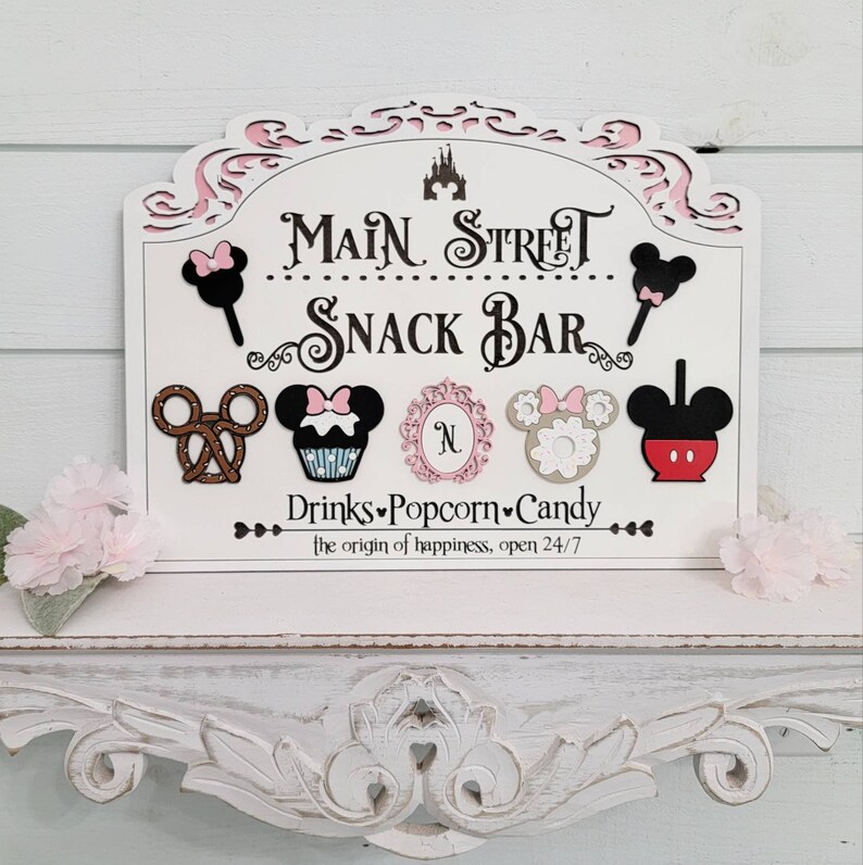 May include: A white wooden sign with pink detailing, reading "Main Street Snack Bar." The sign displays Mickey Mouse-themed snack illustrations, such as ice cream, pretzels, and cupcakes. The text "Drinks, Popcorn, Candy" is also included.