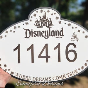 Może przedstawiać: Owalny znak Disneyland Resort z adresem 11416. Znak zawiera grafikę zamku, gwiazdki i frazę "Where Dreams Come True".