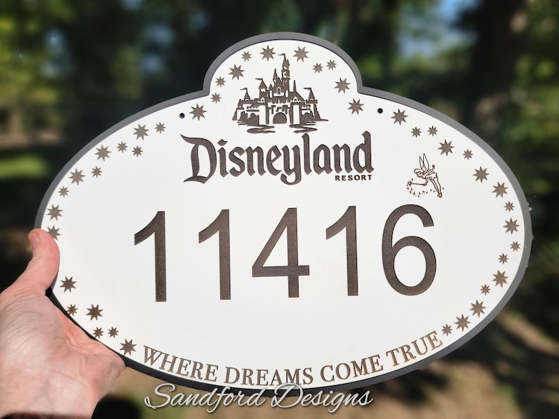 Może przedstawiać: Owalny znak Disneyland Resort z adresem 11416. Znak zawiera grafikę zamku, gwiazdki i frazę "Where Dreams Come True".