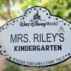 Może przedstawiać: Biały owalny znak z czarną obw&oacute;dką i tekstem. Na znaku widnieje napis "Walt Disney World", "MRS. RILEY'S KINDERGARTEN" i "WHERE DREAMS COME TRUE". Znak ma gwiazdy i grafikę zamku.