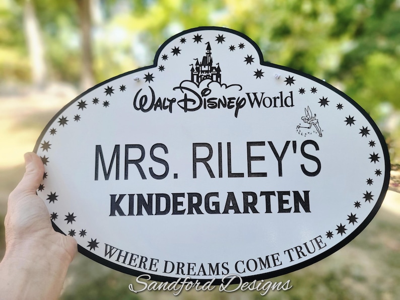 Może przedstawiać: Biały owalny znak z czarną obw&oacute;dką i tekstem. Na znaku widnieje napis "Walt Disney World", "MRS. RILEY'S KINDERGARTEN" i "WHERE DREAMS COME TRUE". Znak ma gwiazdy i grafikę zamku.